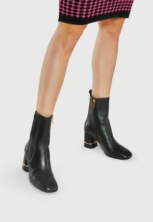 Nogi w czarnych skórzanych botkach za kostkę z złotym zamkiem i słupkowym obcasem, zestawione z różowo-czarną spódnicą w wzory.