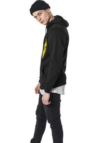 Sudadera con capucha negra y un diseño gráfico amarillo, combinada con jeans oscuros con acentos texturizados. El modelo posa de lado, llevando un gorro.