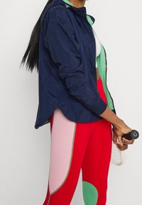 Námořnický větrovka s zelenou podšívkou, s zipovým zapínáním a prodlouženým zadním dílem. V kombinaci s legínami s červeno-růžovým vzorem.