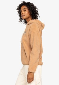 Tan fleece hoodie with a front pocket, white drawstrings, and a small patch logo on the left chest. Curved hem and textured fabric.