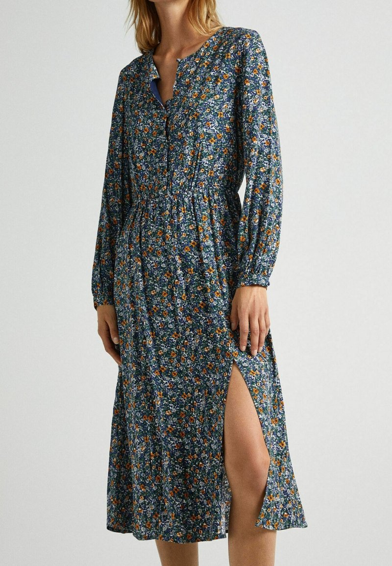 Femme portant une robe midi bleue à fleurs avec manches longues, devant boutonné, taille froncée et une fente haute sur le côté, sur un fond uni.
