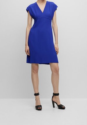 Kvinde iført en knælængde, kortærmet klæde i royalblå med V-hals, kombineret med sorte høje hæle sandaler med ankelstropper.