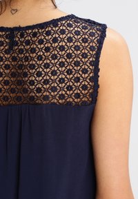 Granatowy top z przezroczystym, haftowanym koronkowym tyłem z kwiatowymi wzorami, uzupełniony gładkim materiałem i falowanymi brzegami.