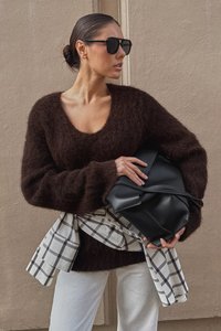 Oversized brown fuzzy sweater with a deep V-neck, paired with a black leather bag and a checked scarf tied around the waist. Light pants.
