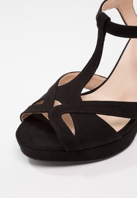 Sandale à talons hauts en daim noir avec bout ouvert, design à bride T et semelle intérieure beige sur fond blanc.