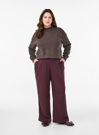 Pull en laine marron à col montant associé à un pantalon large couleur bordeaux. Le modèle a les mains dans les poches et porte des chaussures plates bordeaux.