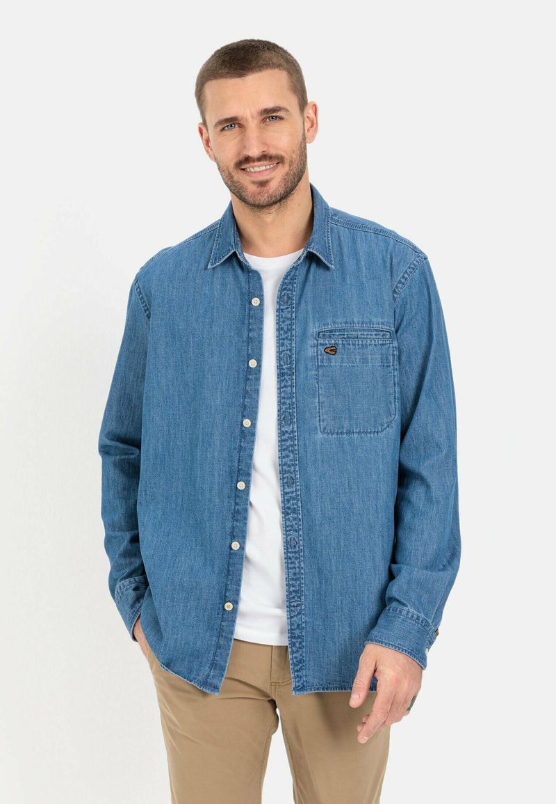 Blaues Denim-Hemd mit Knöpfen und Brusttasche, mit lässiger Passform und langen Ärmeln, über einem weißen T-Shirt getragen.