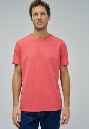 Camiseta corta de manga corta en color coral con un diseño de escote en V y un detalle de logo sutil. Elaborada con una mezcla de algodón texturizado. Combinada con jeans oscuros.