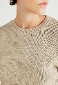 Μπεζ πλεκτό πουλόβερ με στρογγυλή λαιμόκοψη, διαθέτει υφή και ριμπ μανσέτα; το υλικό φαίνεται μαλακό και αναπνεύσιμο.
