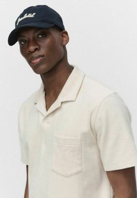 Jasno beżowa koszula z kołnierzykiem, z kieszonką na piersi, wykonana z miękkiego materiału. Noszona z ciemnogranatową czapką z białym haftowanym napisem.