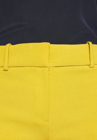 Gele op maat gemaakte broeken van een gladde, gestructureerde stof. Heeft een vlakke tailleband met riemlussen, gestikte details en zijzakken.