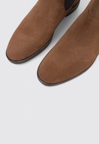 Botas cortas de ante marrón con textura suave, puntera redondeada, paneles elásticos laterales y una suela de goma oscura contrastante.