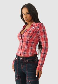 Czerwona koszula w kratę na guziki z długimi rękawami, z białym logo. Czarne spodnie dżinsowe z kontrastującymi szwami i bocznymi kieszeniami.