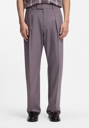 Hombre con pantalones de talle alto, plisados y de pierna recta en color malva, combinados con mocasines de cuero marrón oscuro, de pie frente a un fondo blanco.