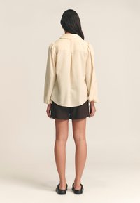 Beige button-up shirt with puff sleeves, curved hemline, and a seam detail at the back, paired with dark shorts and black slip-on shoes.