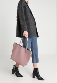 Lichtroze tote bag met een zachte textuur, dubbele handvatten en een open bovenkant. Draag het met een geruite blazer, blauwe cropped jeans en zwarte enkellaarsjes met hak.