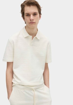 Giovane uomo che indossa una polo a maniche corte color bianco sporco con trama e pantaloni con coulisse abbinati, in piedi con le mani in tasca.