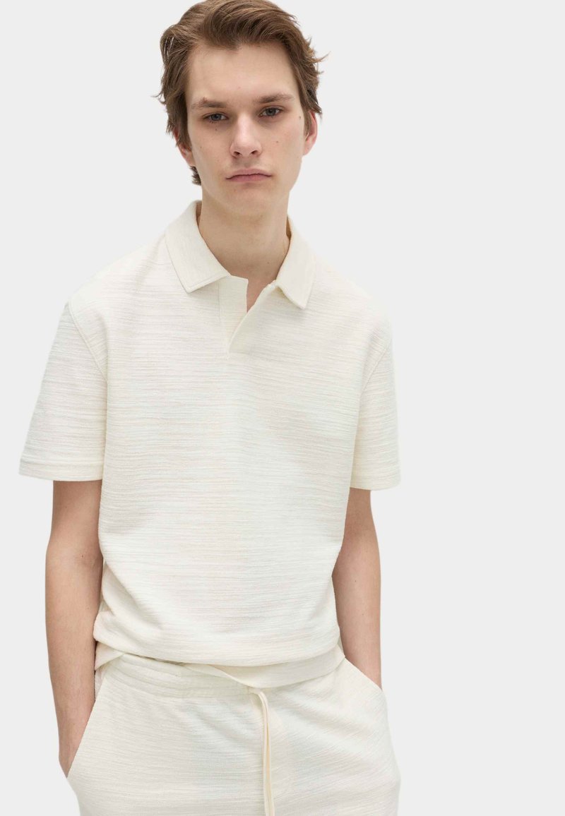 Ung mand iført en tekstureret, off-white polo med korte ærmer og matchende bukser med snøre, stående med hænderne i lommerne.