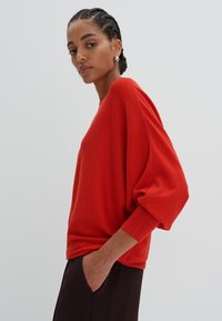 Pull rouge surdimensionné avec des manches longues en ballon, confectionné dans un matériau doux. Poignets et ourlet côtelés, porté sur un pantalon marron foncé.