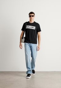 Black cotton t-shirt with "CALVIN KLEIN" logo, light blue jeans, black sneakers, and sunglasses; model carries a small gray crossbody bag.