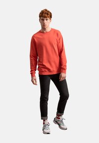 Felpa rossa con maniche lunghe e polsini a coste, abbinata a jeans scuri e sneakers multicolori, su uno sfondo semplice.