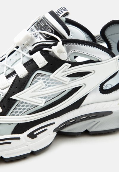 Κοντινό πλάνο μαύρου και λευκού αθλητικού παπουτσιού με πάνελ από δίχτυ, λογότυπο με βέλος και ρυθμιζόμενα ελαστικά κορδόνια με κλείστρο.