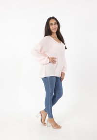 Jasnoróżowy, dziany, oversize'owy sweter z dekoltem w serek, w połączeniu z dopasowanymi niebieskimi dżinsami i przezroczystymi sandałami na obcasie.