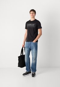 Tricou negru din bumbac cu logo-ul "BARBOUR INTERNATIONAL", asociat cu blugi din denim albastru deschis și o geantă de umă neagră, purtat cu adidași negri.