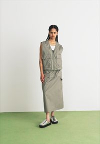 Světlé šedé vesta s velkými kapsami a zipem, spárována s dlouhou ladící sukní. Obuv zahrnuje stříbrné slip-on boty s tlustými černými podrážkami.