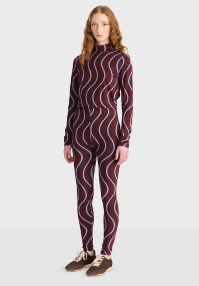 Jeune femme aux longs cheveux roux portant une tenue marron cintrée avec des rayures ondulées bleu clair et des baskets marron, debout devant un fond uni.