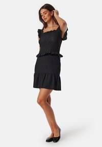Abito in pizzo nero con maniche a ruffle e vita a peplo, caratterizzato da una gonna aderente con orlo svasato. Texture liscia, con motivi floreali visibili.