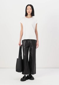 Weißes T-Shirt mit kurzen Ärmeln, schwarze weit geschnittene Lederhosen, schwarze Tote-Tasche mit Tragegurt, schwarze klobige Schuhe, einfarbiger Hintergrund.