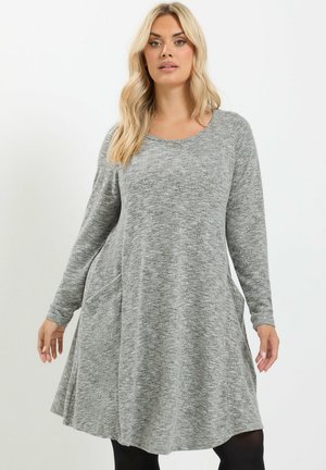 Femme aux longs cheveux blonds portant une robe grise ample à manches longues et des leggings noirs sur un fond clair uni.
