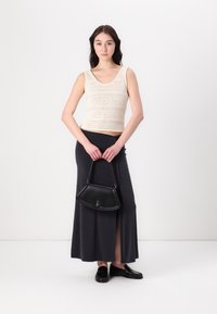 Ljus beige stickad tanktop, svart lång kjol med en sidofriktion, håller en svart läderhandväska och bär svarta loafers.