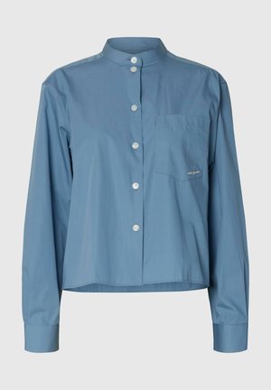 Plava košulja dugih rukava s visokim mandarinskim ovratnikom, gumbima sprijeda, džepom na lijevoj strani prsa i lagano skraćenim rubom.