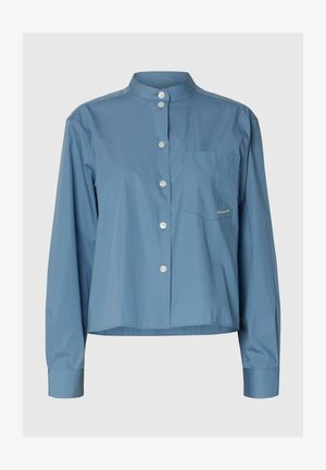 Plava košulja dugih rukava s visokim mandarinskim ovratnikom, gumbima sprijeda, džepom na lijevoj strani prsa i lagano skraćenim rubom.