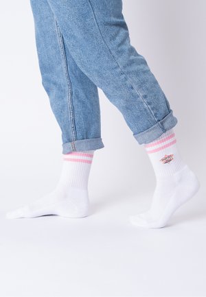 Beine, die hellblaue hochgekrempelte Jeans und weiße Socken mit pinken Streifen und dem Schriftzug „Super Mom“ tragen, gehen auf einer schlichten weißen Oberfläche.