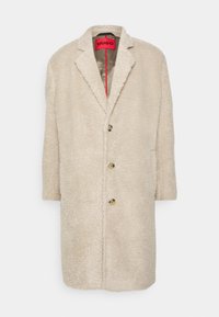 Cappotto beige a mezzaluna in tessuto di lana con colletto a rever, tre bottoni sul davanti e etichetta interna rettangolare rossa con il nome del marchio "HUGO".