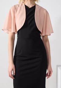 Jasnoróżowe bolerko z szyfonu z falbanowymi rękawami, noszone na dopasowanej czarnej sukience, podkreślające jej miękką fakturę i warstwowy design.