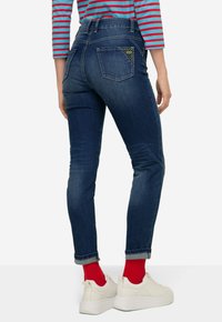 Ciemnoniebieskie dżinsy z wysokim stanem, z unikalnym trójkątnym przeszyciem na tylnej kieszeni, w połączeniu z białymi sneakersami i czerwonymi skarpetkami.