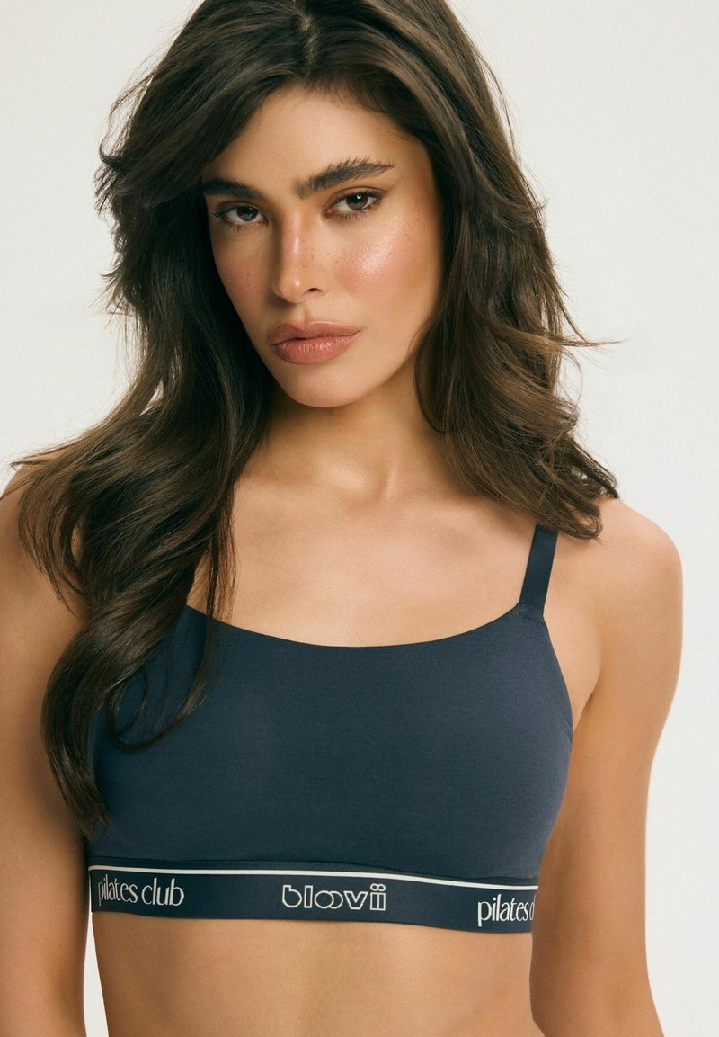 Mladá žena s dlhými hnedými vlasmi, ktorá má na sebe tmavomodrú športovú podprsenku s logom Pilates Club, smerujúca čelom dopredu na jednoduchom pozadí.