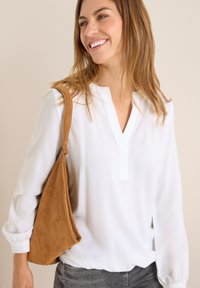 Hvid bluse med V-hals og lange ærmer, parret med en brun ruskind skuldertaske. Blød tekstur, afslappet pasform og enkelt design.