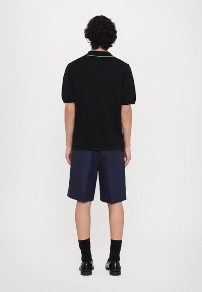 Polo en maille noire à manches courtes avec un motif texturé, assorti à un short bleu marine. Des chaussettes et des chaussures noires complètent la tenue.