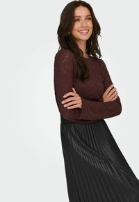 Donna che sorride con le braccia incrociate, indossando un top in pizzo bordeaux a maniche lunghe e una gonna nera plissettata a vita alta su uno sfondo bianco.