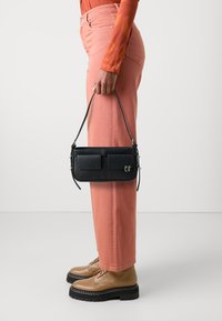 Borsa in pelle nera con tasche frontali, abbinata a pantaloni a gamba larga rosa chiaro e stivali stringati color miele, che evidenziano un design minimalista.