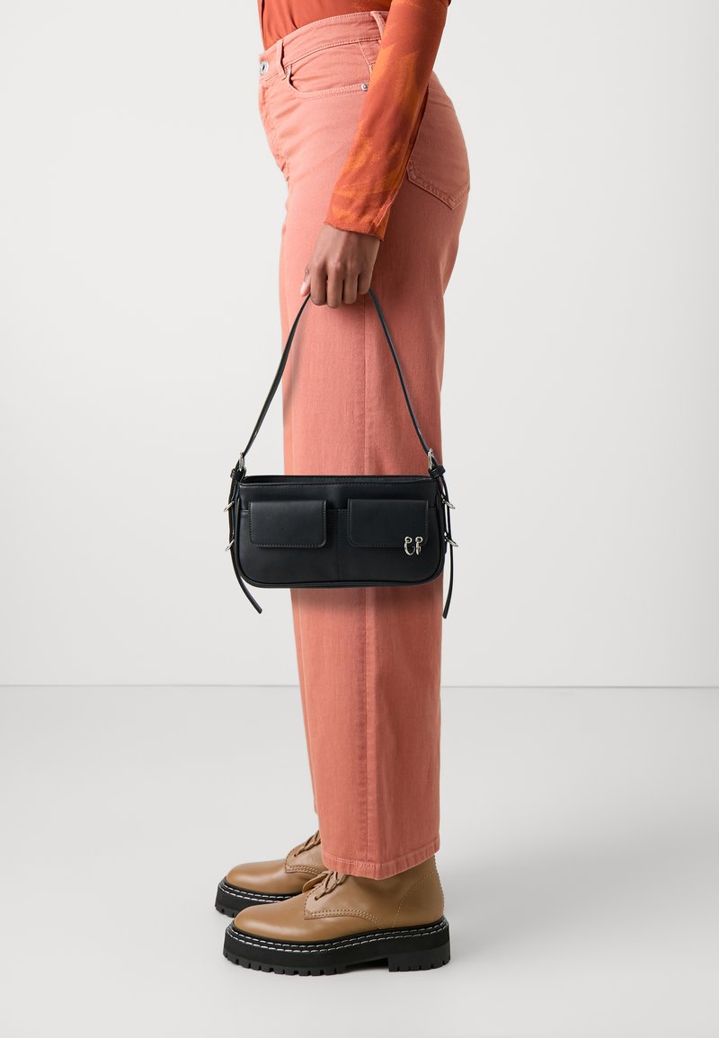 Borsa in pelle nera con tasche frontali, abbinata a pantaloni a gamba larga rosa chiaro e stivali stringati color miele, che evidenziano un design minimalista.