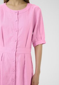 Vestito di lino rosa chiaro con scollo rotondo, maniche corte arrotolate, cinque bottoni sul davanti e vita con cintura.