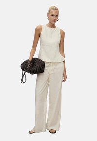 Hellbeige ärmelloses Top mit Peplum-Saum, kombiniert mit weit geschnittenen Hosen. Das Model hält eine schwarze geflochtene Tasche und steht vor einem neutralen Hintergrund.