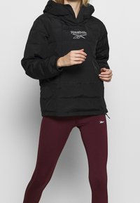 Femme portant une doudoune noire Reebok et des leggings bordeaux Reebok sur un fond clair uni.