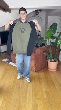 Sudadera con capucha en verde oliva y negro, con el texto "NYC 1990". Corte holgado con un bolsillo frontal. Modelo lleva jeans claros y zapatos plateados.
